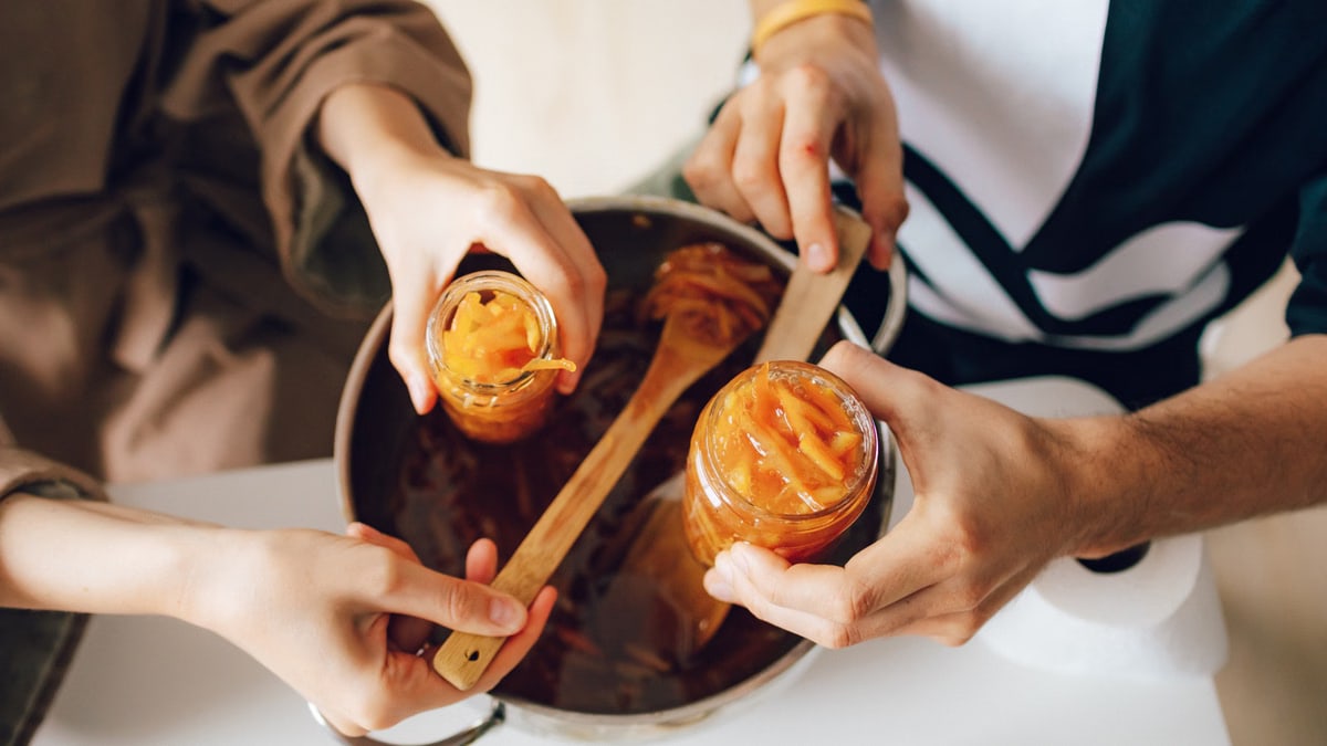 Hands making homemade orange jam, filling jars over a kitchen counter.